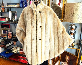 Puede incluir: Un abrigo vintage de piel sintética beige con botones en la parte delantera, exhibido en un maniquí. El abrigo tiene un cuello ancho y un patrón de rayas verticales texturizadas. El maniquí lleva un sombrero marrón y un vestido de encaje negro.
