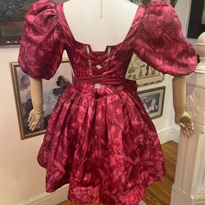 May include: A burgundy dress with a floral pattern, displayed on a mannequin. The dress features a square neckline, puffed sleeves, and a cinched waist with a corset-style back. The skirt is flared, and the fabric has a subtle sheen.