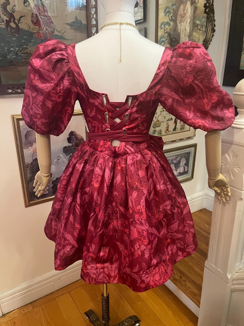 May include: A burgundy dress with a floral pattern, displayed on a mannequin. The dress features a square neckline, puffed sleeves, and a cinched waist with a corset-style back. The skirt is flared, and the fabric has a subtle sheen.