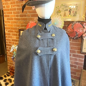 May include: A gray wool cape with gold buttons and the initials "H.M." and "S.A." embroidered on the collar. The cape is paired with a gray hat adorned with feathers. The ensemble is displayed on a mannequin.