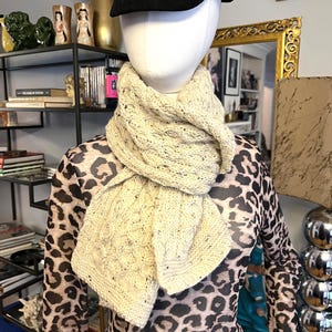 May include: A cream-colored, cable-knit scarf is draped around a mannequin's neck. The scarf has a textured pattern and a slightly speckled appearance. A black cap sits atop the mannequin's head. The mannequin is wearing a long-sleeved leopard print top.