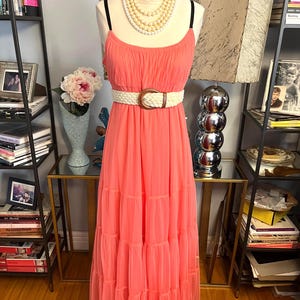 Puede incluir: Vestido largo color coral con tirantes finos y falda escalonada. El vestido se ci&ntilde;e a la cintura con un cintur&oacute;n tejido con una hebilla grande y redonda. Un collar de perlas de varias hebras adorna el cuello del maniqu&iacute;.