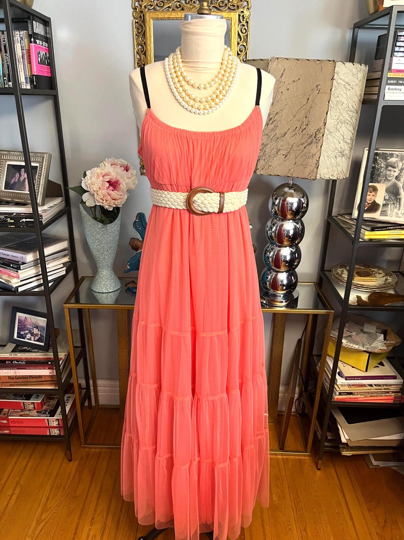 Puede incluir: Vestido largo color coral con tirantes finos y falda escalonada. El vestido se ci&ntilde;e a la cintura con un cintur&oacute;n tejido con una hebilla grande y redonda. Un collar de perlas de varias hebras adorna el cuello del maniqu&iacute;.