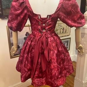 May include: A burgundy dress with a floral pattern, short puff sleeves, and a square neckline. The back features a corset-style lace-up closure and a bow. The dress is displayed on a mannequin.