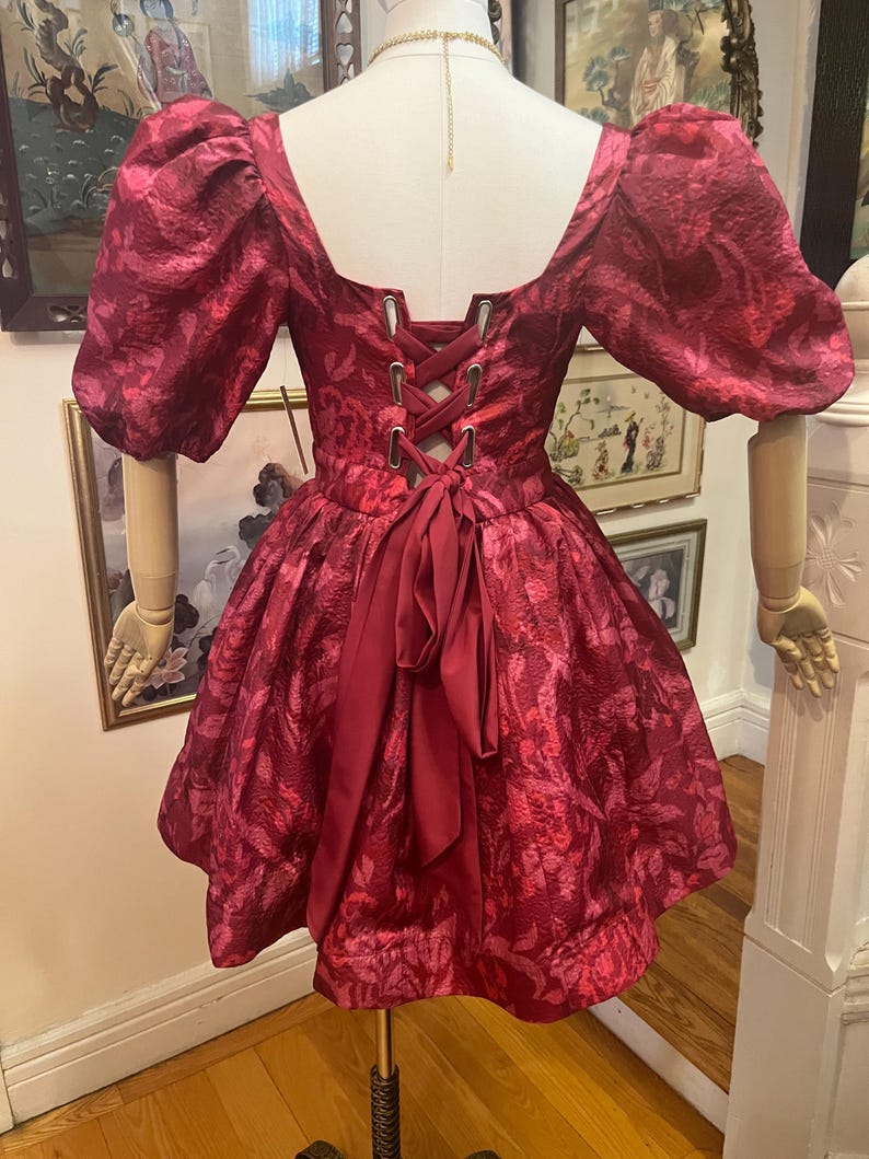 May include: A burgundy dress with a floral pattern, short puff sleeves, and a square neckline. The back features a corset-style lace-up closure and a bow. The dress is displayed on a mannequin.