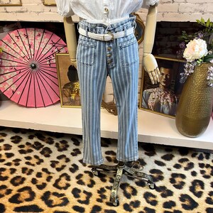 Pode incluir: Uma blusa branca com mangas bufantes e abotoamento &eacute; combinada com jeans listrados em azul e branco. Os jeans t&ecirc;m cintura alta, bolsos frontais e braguilha com bot&otilde;es. Um cinto branco com fivela dourada afivela a cintura.