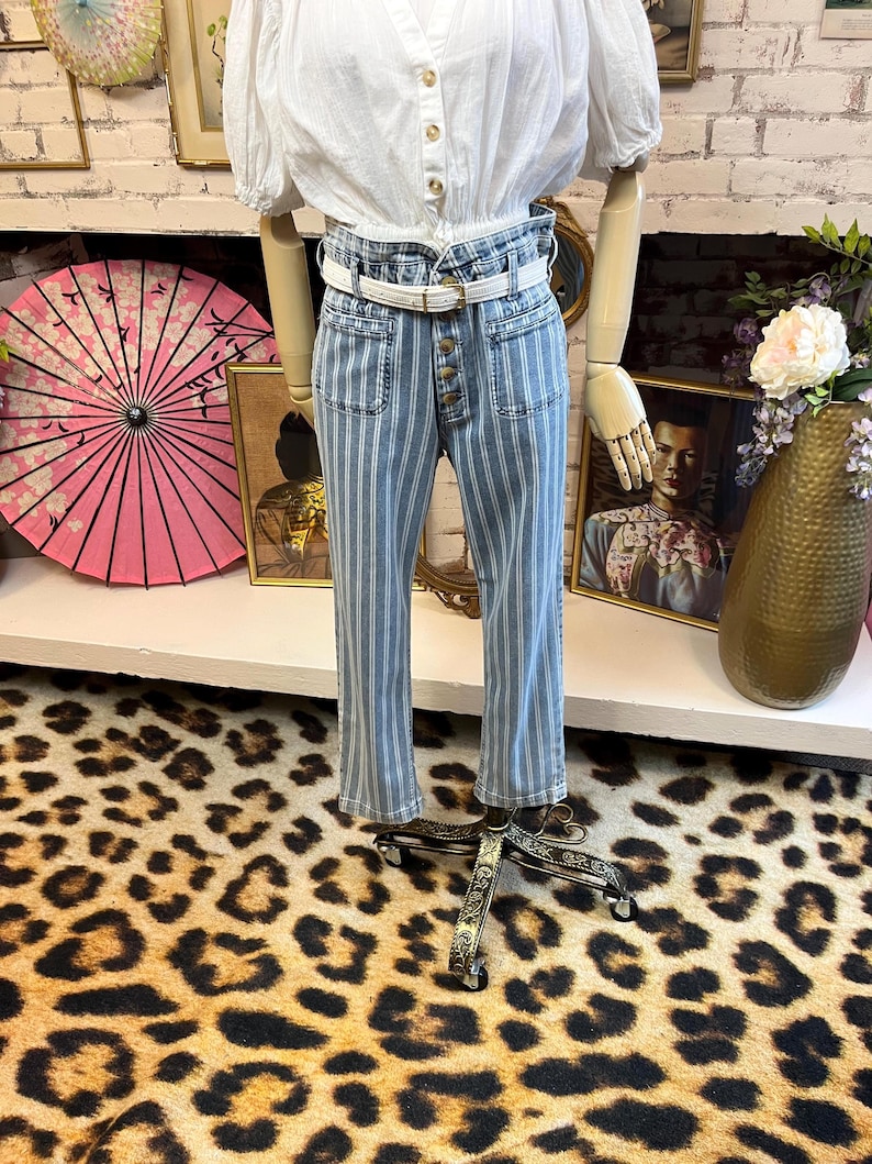 Pode incluir: Uma blusa branca com mangas bufantes e abotoamento &eacute; combinada com jeans listrados em azul e branco. Os jeans t&ecirc;m cintura alta, bolsos frontais e braguilha com bot&otilde;es. Um cinto branco com fivela dourada afivela a cintura.