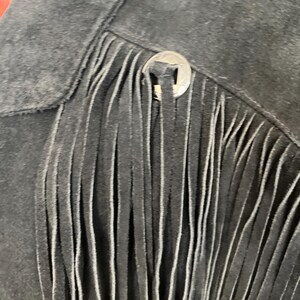 May include: Close-up of a black suede jacket with fringe detailing. The jacket features a silver button closure and a classic western design. The fringe hangs vertically, adding texture and movement to the garment.