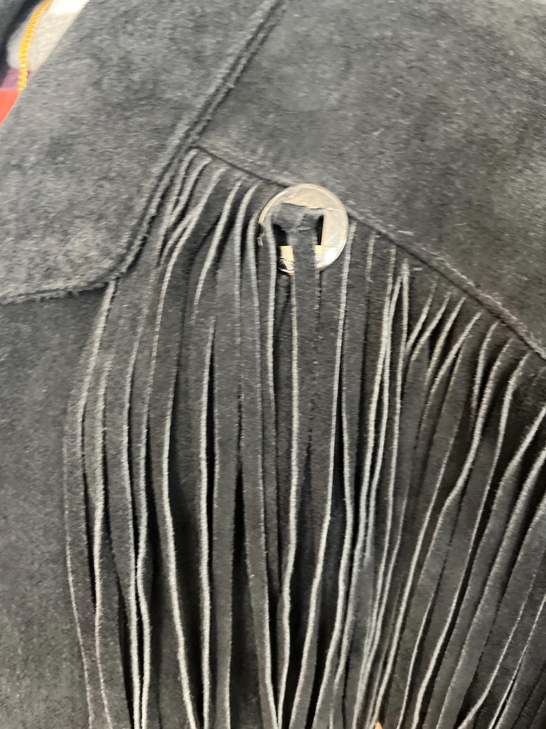 May include: Close-up of a black suede jacket with fringe detailing. The jacket features a silver button closure and a classic western design. The fringe hangs vertically, adding texture and movement to the garment.
