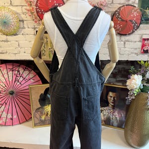 May include: Dark gray denim overalls with adjustable straps and multiple pockets. The overalls are displayed on a mannequin, showcasing the back view of the garment. The denim appears to be pre-washed for a faded look, and the legs are cuffed at the ankles.