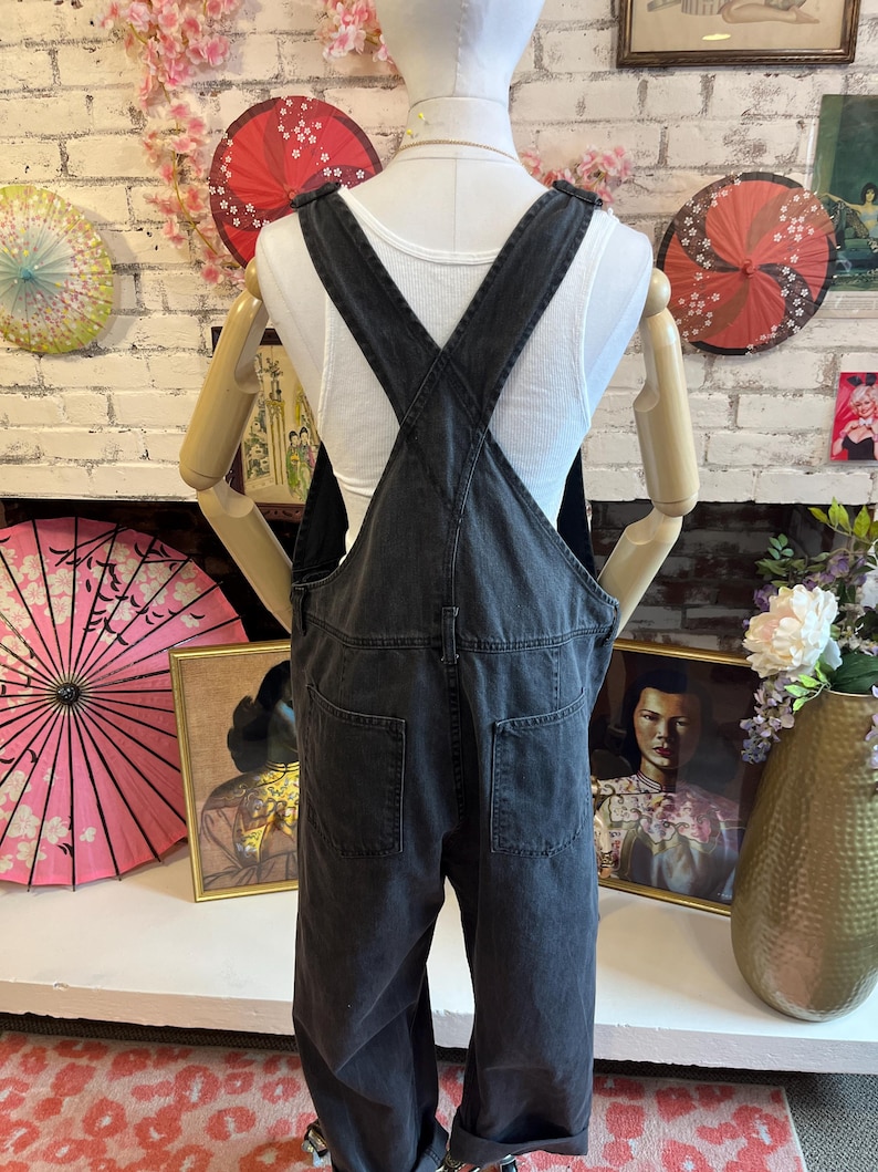 May include: Dark gray denim overalls with adjustable straps and multiple pockets. The overalls are displayed on a mannequin, showcasing the back view of the garment. The denim appears to be pre-washed for a faded look, and the legs are cuffed at the ankles.