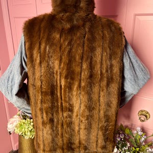 May include: A brown faux fur vest with a high collar. The vest is a rich, warm brown color and has a vertical striped pattern. The vest is worn over a light blue long-sleeved shirt and blue jeans.
