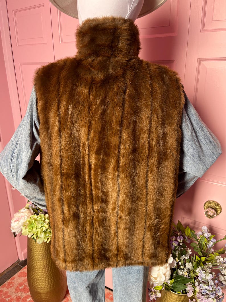 May include: A brown faux fur vest with a high collar. The vest is a rich, warm brown color and has a vertical striped pattern. The vest is worn over a light blue long-sleeved shirt and blue jeans.