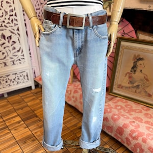 May include: Light blue denim jeans featuring a distressed finish with small frayed areas on the legs. The jeans are styled with a brown leather belt that has a large, square, silver-toned metal buckle. The pant legs are cuffed at the bottom hem.