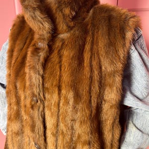 May include: A vintage brown fur vest with a high collar and button closure. The vest is layered over a light blue denim jacket. The fur has a rich, reddish-brown color and a soft, textured appearance. The vest appears to be in good condition.