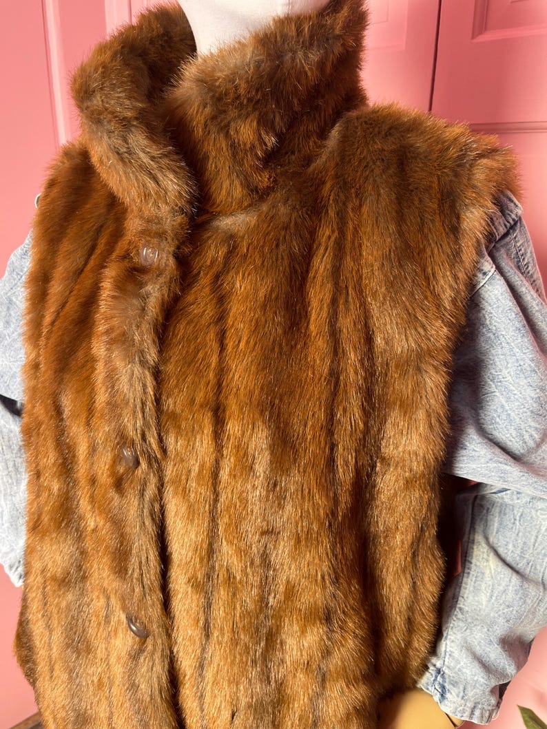 May include: A vintage brown fur vest with a high collar and button closure. The vest is layered over a light blue denim jacket. The fur has a rich, reddish-brown color and a soft, textured appearance. The vest appears to be in good condition.