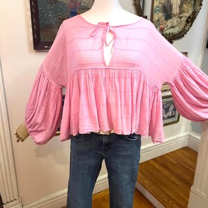 May include: A light pink blouse with long, voluminous sleeves and a gathered bodice. The top features a keyhole neckline with a tie closure. The blouse is paired with blue denim jeans.