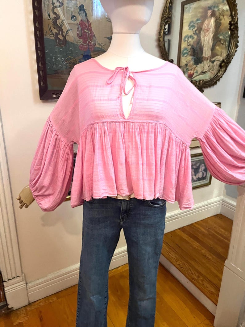 May include: A light pink blouse with long, voluminous sleeves and a gathered bodice. The top features a keyhole neckline with a tie closure. The blouse is paired with blue denim jeans.