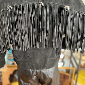 May include: Black suede jacket with fringe detail across the back. The jacket features silver-toned button accents and a cropped silhouette. The fringe hangs vertically, adding a Western-inspired flair to the garment.