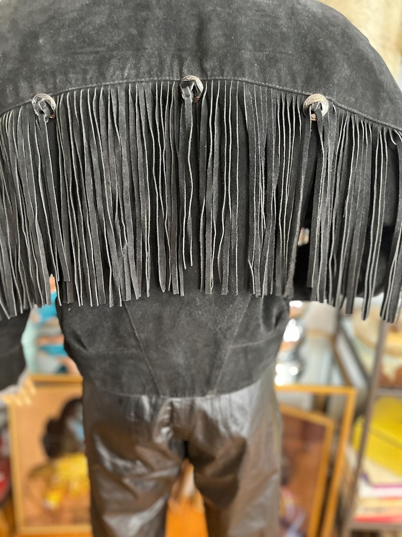 May include: Black suede jacket with fringe detail across the back. The jacket features silver-toned button accents and a cropped silhouette. The fringe hangs vertically, adding a Western-inspired flair to the garment.