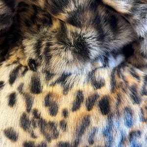 K&ouml;nnte beinhalten: Eine Nahaufnahme eines weichen, strukturierten Stoffes mit einem markanten Leopardenmuster. Das Material zeigt eine Mischung aus beigen, goldbraunen und schwarzen fellartigen Fasern, die ein dichtes, geschichtetes Aussehen mit dunklen Flecken erzeugen.