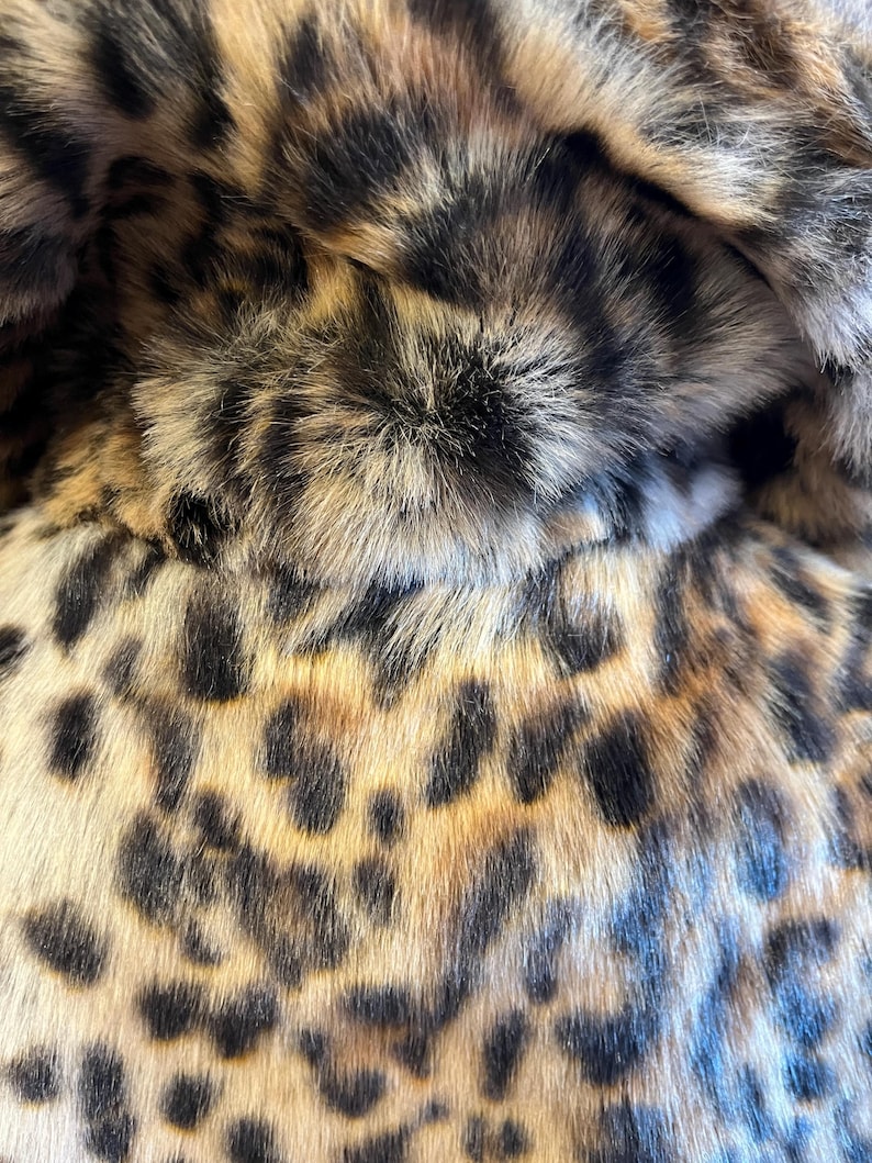 K&ouml;nnte beinhalten: Eine Nahaufnahme eines weichen, strukturierten Stoffes mit einem markanten Leopardenmuster. Das Material zeigt eine Mischung aus beigen, goldbraunen und schwarzen fellartigen Fasern, die ein dichtes, geschichtetes Aussehen mit dunklen Flecken erzeugen.