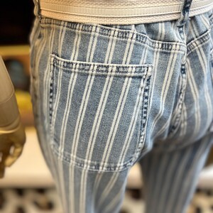 Pode incluir: Close-up de cal&ccedil;as de ganga azul claro com riscas brancas verticais. As cal&ccedil;as t&ecirc;m um cinto branco e um bolso traseiro. O tecido &eacute; ganga clara. As cal&ccedil;as fazem parte de uma cole&ccedil;&atilde;o de roupa casual.