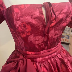 May include: A formal, floor-length dress in a deep red hue with a floral pattern. The dress features a square neckline, a cinched waist with a matching sash, and a large bow. The fabric has a subtle sheen.