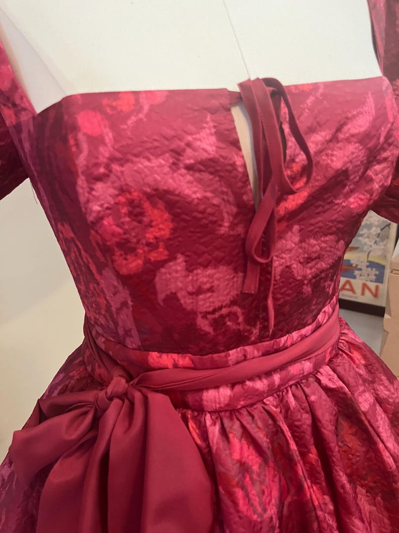 May include: A formal, floor-length dress in a deep red hue with a floral pattern. The dress features a square neckline, a cinched waist with a matching sash, and a large bow. The fabric has a subtle sheen.