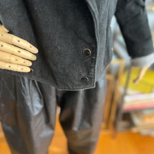 May include: Black suede jacket with fringe detail on the sleeves. The jacket has a rounded collar and two visible buttons. The model is wearing black leather pants. The jacket is a vintage style.
