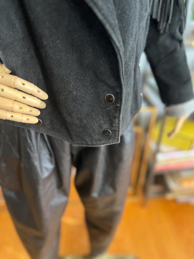 May include: Black suede jacket with fringe detail on the sleeves. The jacket has a rounded collar and two visible buttons. The model is wearing black leather pants. The jacket is a vintage style.