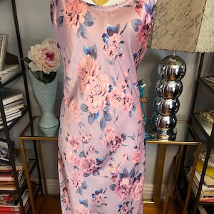 May include: A lilac-colored slip dress with a floral pattern of pink and blue roses. The dress has thin spaghetti straps and a gold pendant necklace. The dress is displayed on a mannequin.