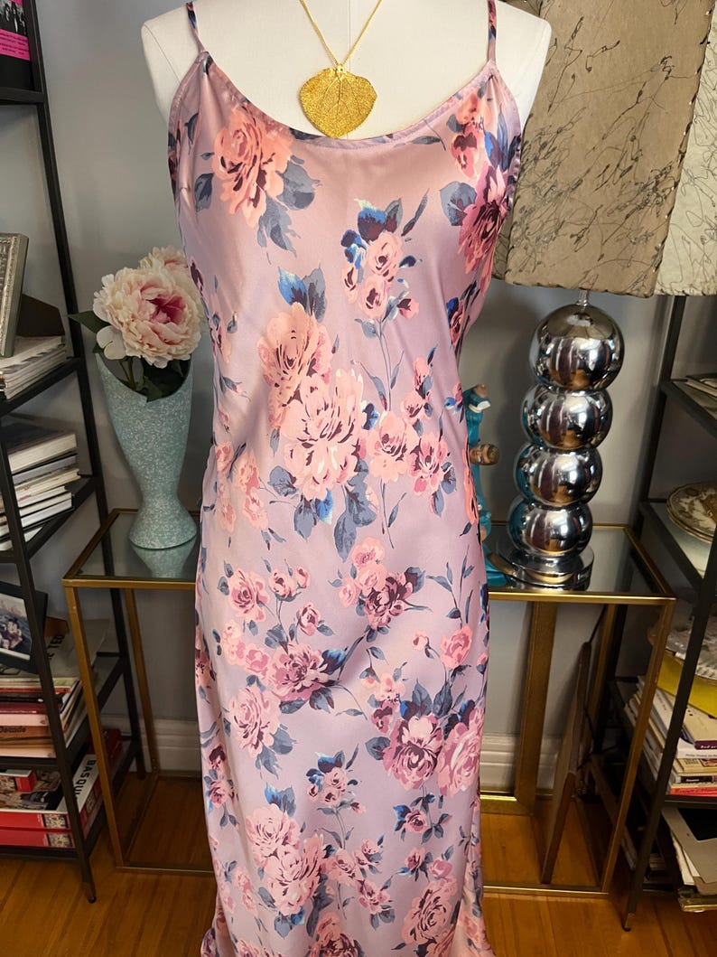May include: A lilac-colored slip dress with a floral pattern of pink and blue roses. The dress has thin spaghetti straps and a gold pendant necklace. The dress is displayed on a mannequin.