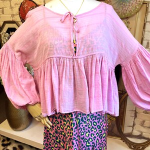 May include: A light pink blouse with long, voluminous sleeves and a gathered bodice. The blouse has a tie detail at the neckline. The mannequin is also wearing a pink and green leopard print skirt. The blouse is displayed on a mannequin.