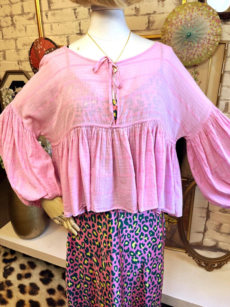 May include: A light pink blouse with long, voluminous sleeves and a gathered bodice. The blouse has a tie detail at the neckline. The mannequin is also wearing a pink and green leopard print skirt. The blouse is displayed on a mannequin.