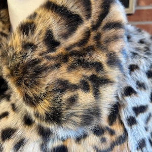 K&ouml;nnte beinhalten: Ein Mantel aus dichtem, strukturiertem Kunstfell mit einem markanten Leopardenmuster. Das Design zeigt eine Mischung aus hellbraunen, dunkelbraunen und schwarzen Flecken auf der Oberfl&auml;che, mit einem gro&szlig;en, umgeschlagenen Kragen im oberen Bereich.