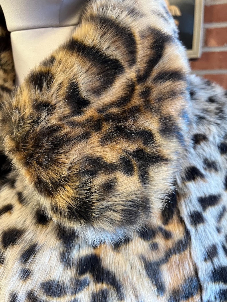 K&ouml;nnte beinhalten: Ein Mantel aus dichtem, strukturiertem Kunstfell mit einem markanten Leopardenmuster. Das Design zeigt eine Mischung aus hellbraunen, dunkelbraunen und schwarzen Flecken auf der Oberfl&auml;che, mit einem gro&szlig;en, umgeschlagenen Kragen im oberen Bereich.