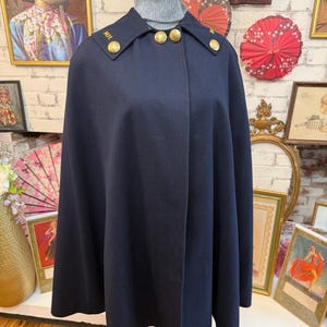 May include: A dark navy blue wool-blend cape featuring a high collar, a grey turtleneck sweater underneath, and gold-toned metal buttons on the collar points and front closure. The collar points are embroidered with the letters M.I. and M.A.