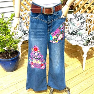 May include: Blue denim wide-leg jeans with colorful floral embroidery on the front and pocket. The jeans have a brown leather belt with a silver buckle. The jeans are displayed on a mannequin.