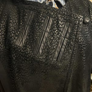 May include: Black leather jacket with a spotted pattern. The jacket has a notched collar, three vertical accent panels, and a single button closure. The jacket is layered over a denim jacket and a patterned scarf.