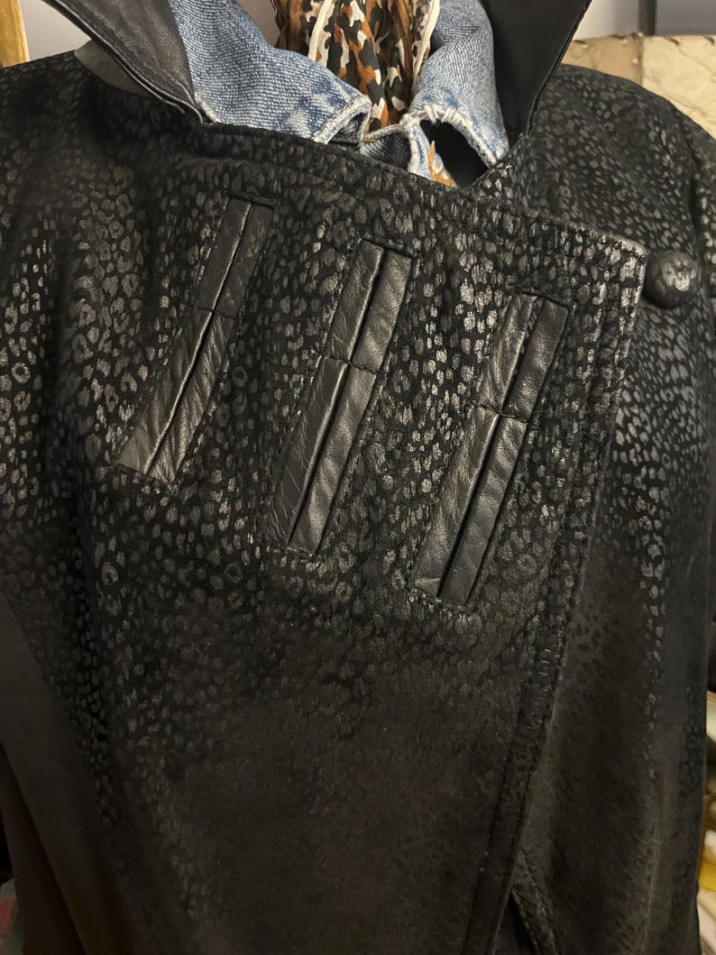 May include: Black leather jacket with a spotted pattern. The jacket has a notched collar, three vertical accent panels, and a single button closure. The jacket is layered over a denim jacket and a patterned scarf.
