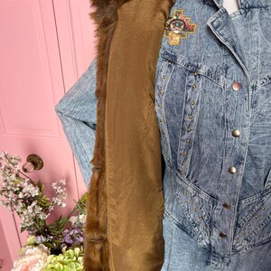 May include: A vintage denim jacket with a brown fur collar and lining. The jacket features gold-colored stud accents and a decorative pin with turquoise and colorful details. The jacket is displayed on a mannequin.