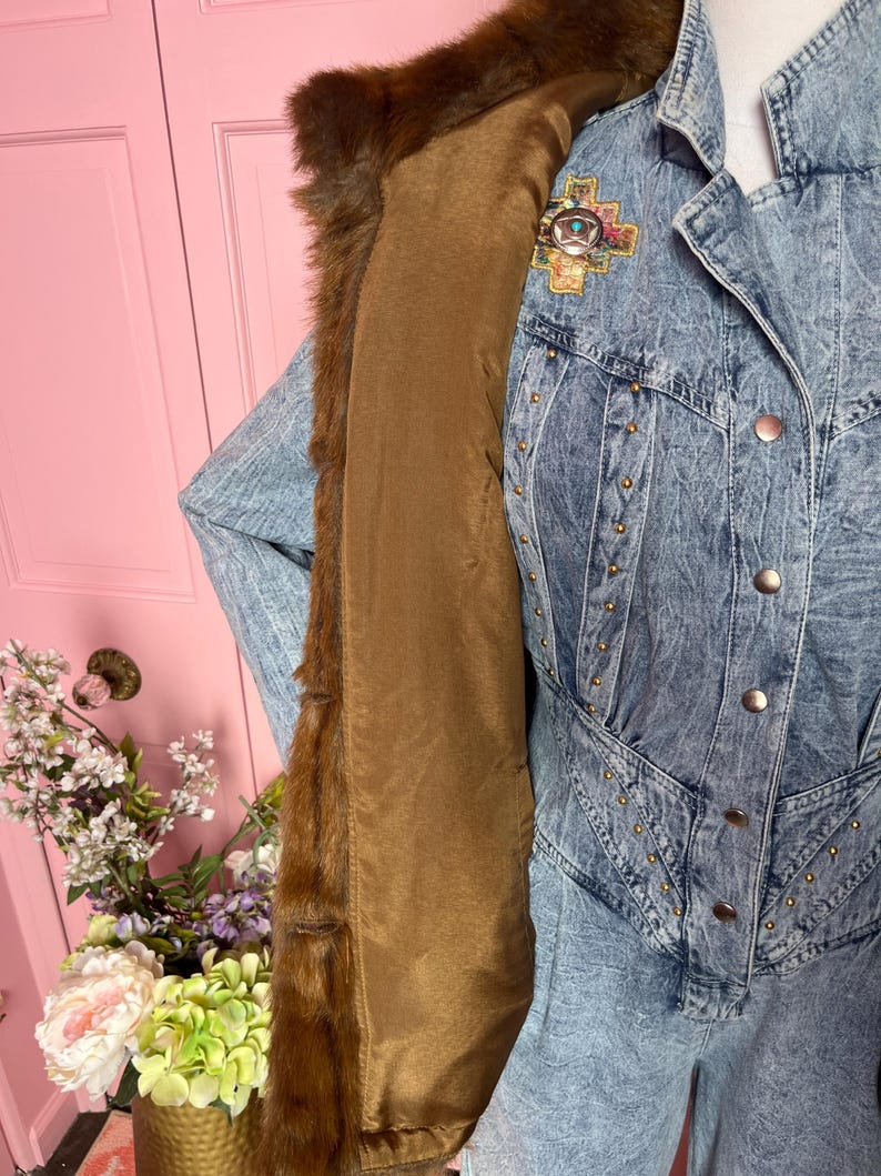May include: A vintage denim jacket with a brown fur collar and lining. The jacket features gold-colored stud accents and a decorative pin with turquoise and colorful details. The jacket is displayed on a mannequin.