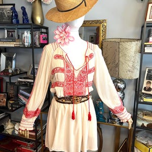 May include: A cream-coloured dress with red embroidery and long sleeves is displayed on a mannequin. The dress features a V-neck, tassels, and a patterned belt. A straw hat and a pink flower adorn the mannequin's head and neck, respectively.