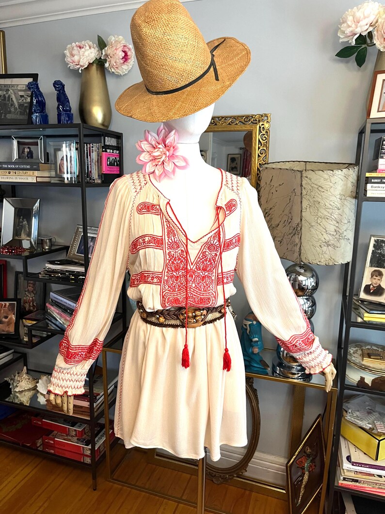May include: A cream-coloured dress with red embroidery and long sleeves is displayed on a mannequin. The dress features a V-neck, tassels, and a patterned belt. A straw hat and a pink flower adorn the mannequin's head and neck, respectively.
