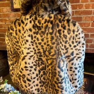 K&ouml;nnte beinhalten: Eine kurze Jacke mit einem markanten Leopardenmuster in Beige-, Braun- und Schwarzt&ouml;nen. Das Kleidungsst&uuml;ck hat einen breiten, umgeschlagenen Kragen und eine dicke, strukturierte Kunstfelloptik, getragen &uuml;ber einer hellblauen Jeans.