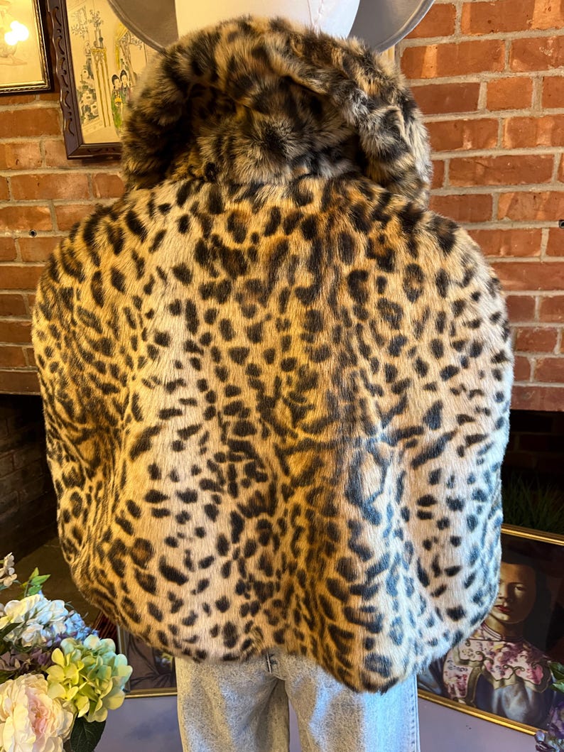 K&ouml;nnte beinhalten: Eine kurze Jacke mit einem markanten Leopardenmuster in Beige-, Braun- und Schwarzt&ouml;nen. Das Kleidungsst&uuml;ck hat einen breiten, umgeschlagenen Kragen und eine dicke, strukturierte Kunstfelloptik, getragen &uuml;ber einer hellblauen Jeans.
