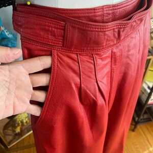 May include: Close-up of red leather pants with a high waist and pleated front. The pants have a wide belt and pockets. The leather appears to be soft and has a slight sheen. The pants are displayed on a mannequin.