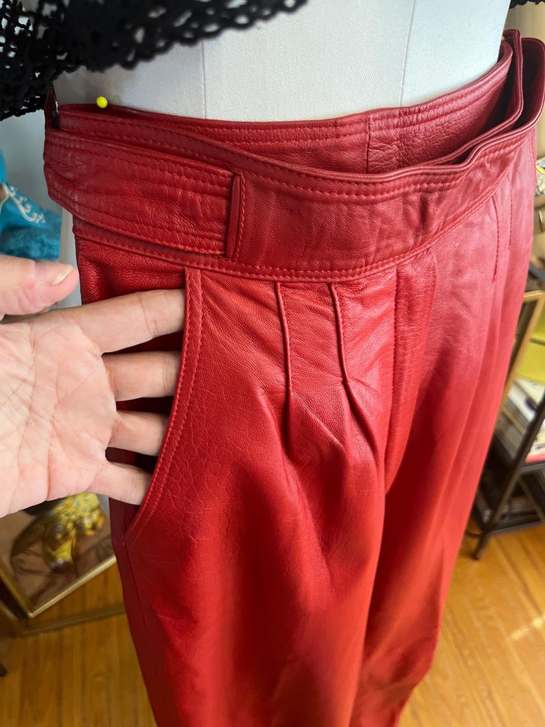 May include: Close-up of red leather pants with a high waist and pleated front. The pants have a wide belt and pockets. The leather appears to be soft and has a slight sheen. The pants are displayed on a mannequin.