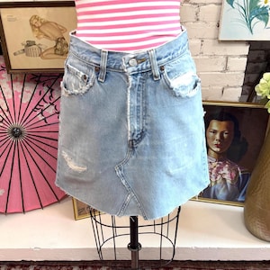 May include: A light blue denim mini skirt featuring a high-waisted design, a button and zipper fly closure, distressed frayed edges at the pockets, and a raw hemline. The skirt has a visible diagonal seam detail across the front panel.